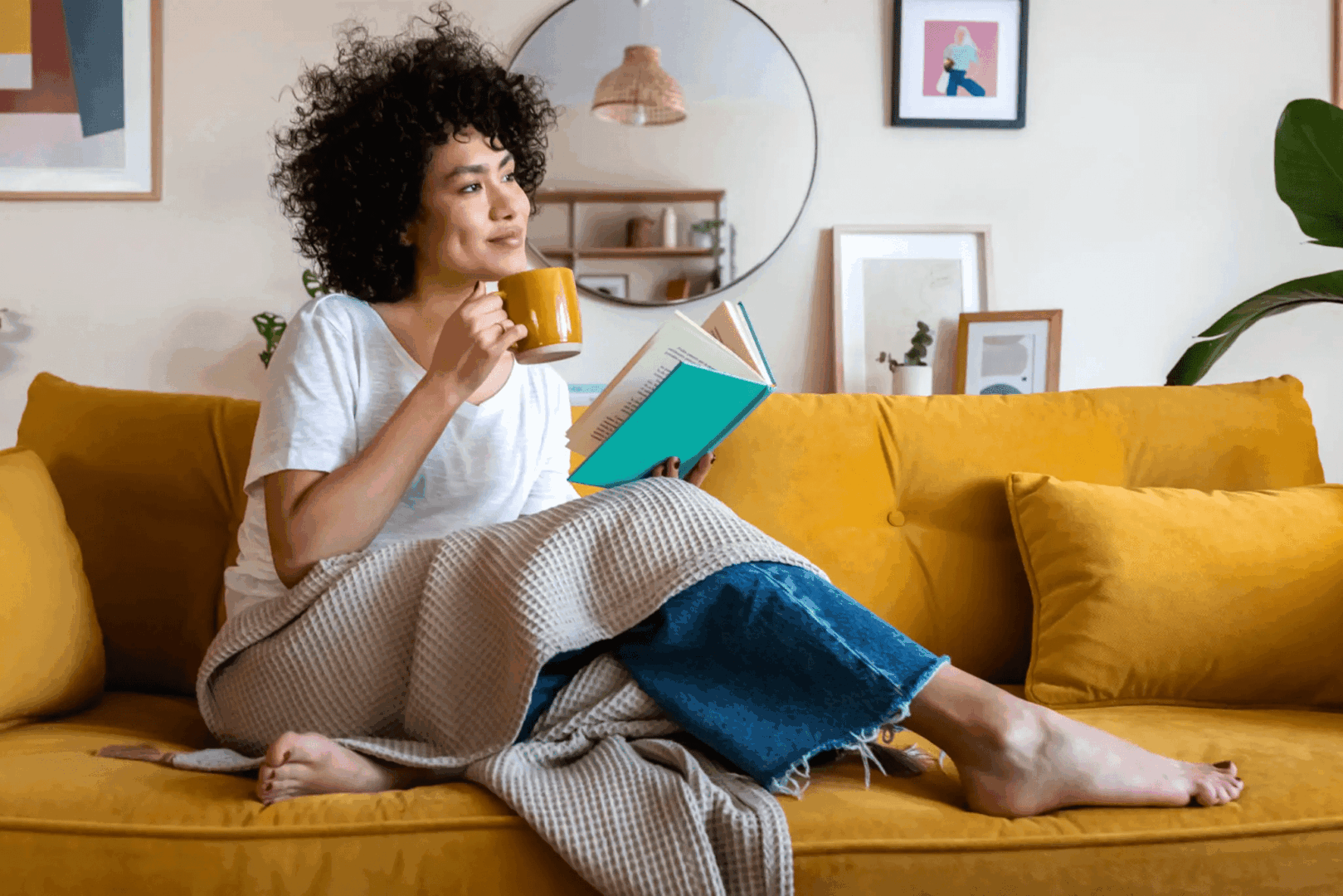 A woman enjoys coffee while reading a book on a yellow couch.