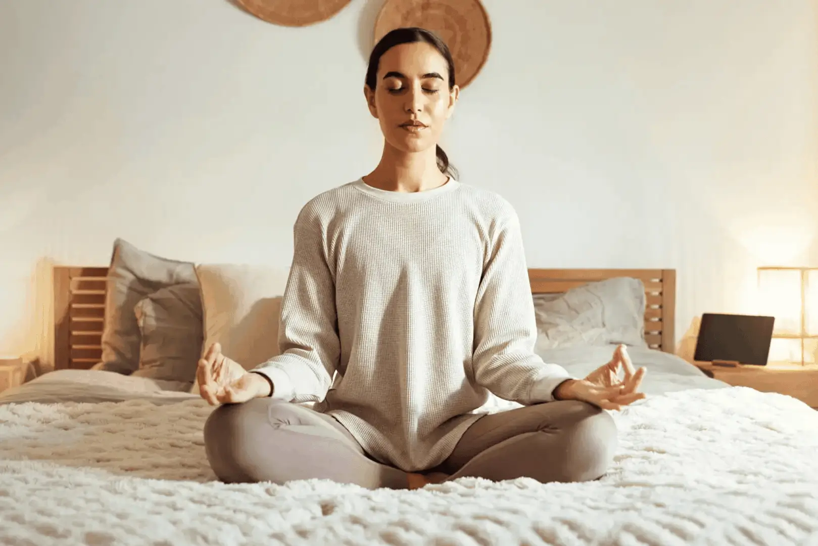 A woman meditates calmly on a bed in a cozy room.